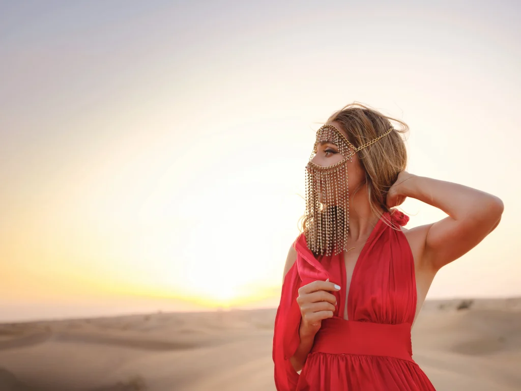 A woman with blonde hair wearing a red dress and a gold, beaded Arabian face veil posing in the sandy Dubai desert as the sun sets on the horizon.