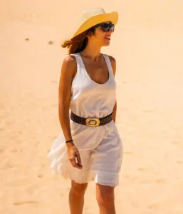 A woman in a white dress, sunglasses, and a hat walking across the undulating sand dunes under a bright sky.