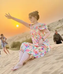 A woman in a colorful dress reaching out towards the setting sun over the rolling sand dunes.