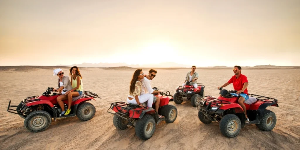 Group of six peoples riding red quad bikes in a desert during sunset, enjoying a desert safari adventure with clear skies and distant mountains in the background.