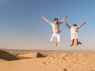 Happy couple jumping in the golden sands of Dubai desert during a Desert Safari adventure under a clear blue sky – perfect for booking unforgettable Desert Safari Dubai tickets.